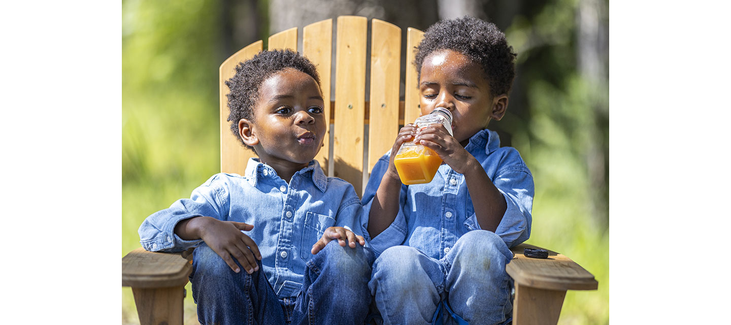 Twin boys drinking orange juice while sitting together on a wooden chair.